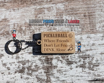Puede incluir: Un llavero de madera con fondo marrón y letras doradas. El llavero presenta un gráfico de una pala y una pelota de pickleball y el texto "Pickleball Where Friends Don't Let Friends DINK Alone."
