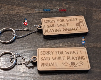 Puede incluir: Dos llaveros de madera con una cadena plateada. Los llaveros son rectangulares y tienen el texto "Lo siento por lo que dije mientras jugaba al pinball". Un llavero tiene un gráfico de máquina de pinball y el otro tiene un gráfico de flipper de pinball.
