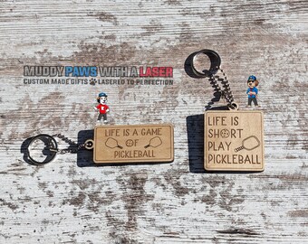 Puede incluir: Dos llaveros de madera con los textos "LIFE IS A GAME OF PICKLEBALL" y "LIFE IS SHORT PLAY PICKLEBALL". Cada llavero tiene un anillo y una cadena de color plateado. Los llaveros son de color marrón claro con diseños grabados de palas y pelotas de pickleball.