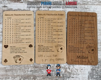 Puede incluir: Tres cartas de juego de madera con el título "Absolute Frustration Rummy" y una lista de reglas para el juego. Las cartas son de color marrón claro con texto negro y una ilustración en blanco y negro de cartas de juego. La carta inferior tiene el texto "Miller Family Game Night".