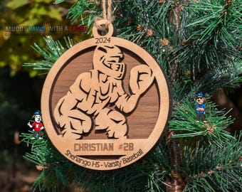Puede incluir: Un adorno navideño de madera con un jugador de béisbol en posición de receptor. El adorno es de color marrón con un contorno negro y el texto "2024", "CHRISTIAN #28" y "Shenango HS - Varsity Baseball".