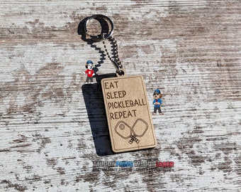 Puede incluir: Llavero de madera con fondo marrón y el texto "EAT SLEEP PICKLEBALL REPEAT" con un dibujo de dos palas de pádel.