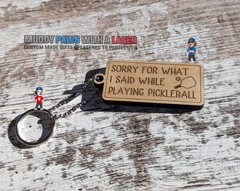 Puede incluir: Un llavero de madera con fondo marrón y texto blanco que dice "Lo siento por lo que dije mientras jugaba al pickleball". Una pequeña bola blanca y una pala de pickleball en blanco y negro están ilustradas en el llavero.