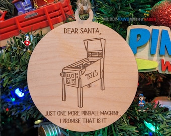 Puede incluir: Un adorno navideño de madera con un diseño de máquina de pinball grabado con láser. El adorno dice "Querido Santa, Solo una máquina de pinball más, te lo prometo, es todo." y presenta el año 2023.