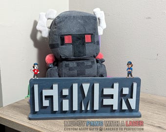 Puede incluir: Un letrero de madera gris con la palabra "GAMER" en letras blancas. Un personaje de peluche estilo Minecraft está detrás del letrero, con dos pequeñas figuras encima. El letrero está sobre una superficie de madera.
