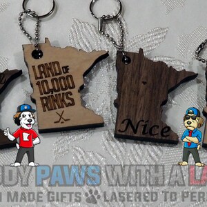 May include: Four wooden keychains shaped like the state of Minnesota. Each keychain has a different inscription: "Home", "Land of 10,000 Rinks", "Nice", and "Duck Duck Gray Duck!"