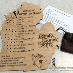 May include: Wooden game board shaped like the state of Michigan with the words "Absolute Frustration Rummy" and "Family Game Night" engraved on it. The board has a list of rules for the game and a set of pegs for keeping score. The board is accompanied by a brown velvet bag and a set of playing cards.