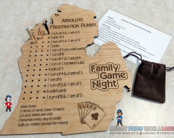 Puede incluir: Un tablero de juego de madera con forma del estado de Michigan con las palabras "Absolute Frustration Rummy" y "Family Game Night" grabadas en él. El tablero tiene una lista de reglas para el juego y un conjunto de clavijas para llevar la puntuación. El tablero viene acompañado de una bolsa de terciopelo marrón y un juego de cartas.
