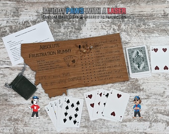 Puede incluir: Un tablero de juego de madera con forma del estado de Montana, con las palabras "Absolute Frustration Rummy" y una lista de reglas para el juego. Hay dos barajas de cartas, una bolsa verde y dos perros de dibujos animados en la mesa.