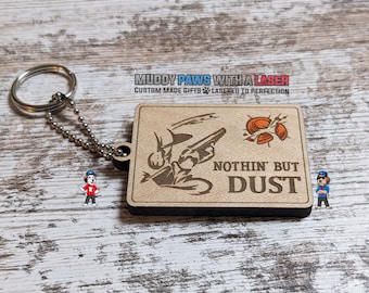 Puede incluir: Un llavero de madera con un diseño grabado con láser de un plato de arcilla siendo disparado. El texto "NOTHIN' BUT DUST" también está grabado en el llavero.