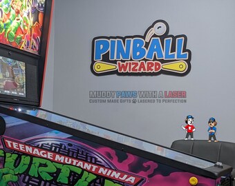 Puede incluir: Una máquina de pinball de las Tortugas Ninja con un campo de juego colorido. Arriba, un letrero dice "Pinball Wizard" en azul y amarillo. Dos figuras de perros están cerca. La pared también muestra el texto "MUDDY PAWS WITH A LASER".