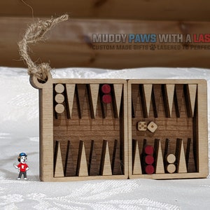 May include: A wooden backgammon game ornament with red and white playing pieces. The ornament is in the shape of a small box with a hinged lid. The ornament is made by Muddy Paws With A Laser, a company that makes custom made gifts.