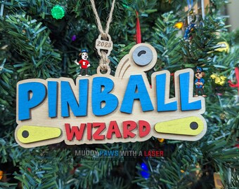 Puede incluir: Un adorno navideño de madera con las palabras "Pinball Wizard" en letras azules y rojas. El adorno está decorado con un flipper amarillo y negro y una bola de pinball gris. El adorno está fechado en 2023.
