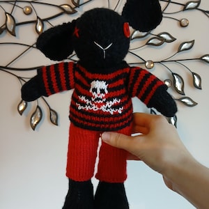 May include: A handmade black knitted rabbit plush toy with long ears, one red button eye, and one stitched red cross eye. It wears a red and black striped jumper featuring a white skull and crossbones graphic, paired with solid red knitted trousers.