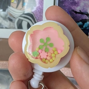 Könnte beinhalten: Ein weißes, einziehbares Ausweis-Jojo mit einem transparenten Kunststoffband. Das Jojo hat ein blütenförmiges Design mit rosa und gelben Konfetti im Inneren. Der Hintergrund umfasst gerahmte Kunstwerke und dekorative Gegenstände.