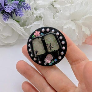 May include: A black circular phone grip with two popsicle-shaped designs. The popsicles are light green with glitter and small black details. The grip is decorated with pink flowers, iridescent beads, and small pearls.