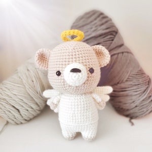 May include: A crocheted teddy bear wearing a halo and angel wings. The bear is light brown with a white face and white wings. The halo is yellow.