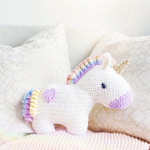 Puede incluir: Un unicornio de peluche blanco tejido a crochet con una melena y cola arcoíris. El unicornio tiene un corazón morado en un lado y está sentado en una almohada con estampado blanco y beige.