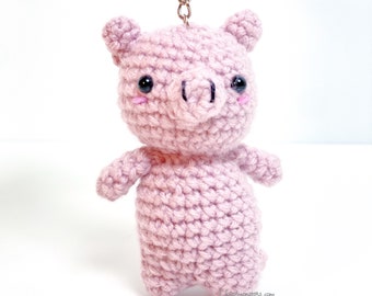 May include: A handmade, light pink crochet pig keychain. The pig has black eyes, a pink snout, and rosy cheeks. The keychain is attached to a silver chain. The pig is standing on a white surface.