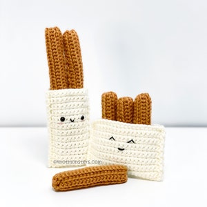 May include: Three crocheted churros in a white bag with a smiling face. Two more churros are in a smaller white bag with a smiling face. A single crocheted churro is lying on the white surface.  ©knotmonsters.com