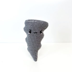 May include: A gray crocheted tornado with a cute cartoon face. The tornado has black eyes and a frown, and pink cheeks.