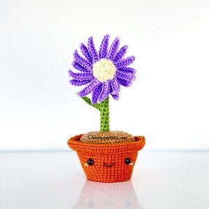 May include: A crocheted purple flower with a yellow center is in a small orange pot with a happy face. The pot is sitting on a white surface. ©knotmonsters.com