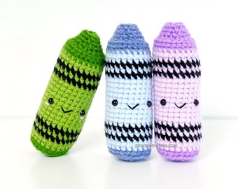 May include: Three crocheted crayon toys with smiling faces. The crayons are green, blue, and purple with black stripes.