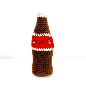 Peut inclure: Une bouteille de soda en crochet marron avec une étiquette rouge et blanche et un visage souriant. La bouteille est posée sur une surface blanche.
