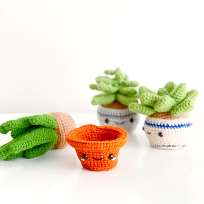 May include: Three crocheted succulent plants in pots with happy faces. The pots are orange, grey and white with blue stripes.  The plants are green and have a 3D appearance.  The text "knotmonsters.com" is visible in the bottom right corner.