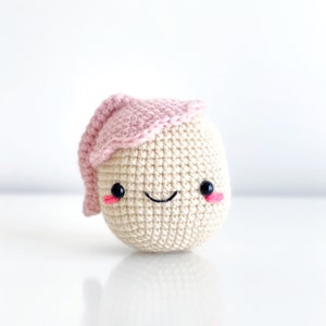 May include: A crocheted plush toy shaped like an egg with a pink crocheted hat. The toy has a smiling face with black eyes and pink cheeks.