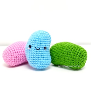 Pode incluir: Três peluches de crochê em forma de feijão. Um é rosa, outro é azul com uma carinha feliz e o último é verde.