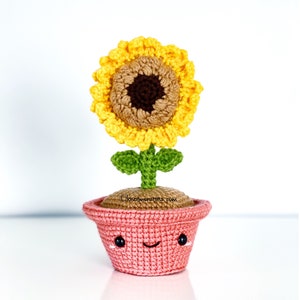 May include: A crocheted sunflower in a pink pot with a smiling face. The sunflower has yellow petals, a brown centre, and green leaves and stem. The pot is a light pink colour.