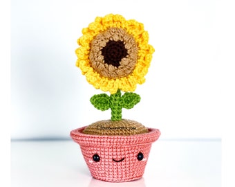 Op de afbeelding: Een gehaakte zonnebloem in een roze pot met een lachend gezicht. De zonnebloem heeft gele bloemblaadjes, een bruin midden en groene bladeren en stengel. De pot is lichtroze.