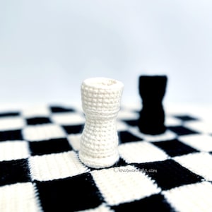 May include: A crocheted white chess rook stands on a black and white checkered surface, with a black rook in the background. The rook is cylindrical with a flared top. The background is a soft white.