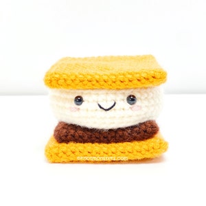 May include: A crocheted s'more plush toy with a smiling face. The s'more is made of brown, white, and yellow yarn.