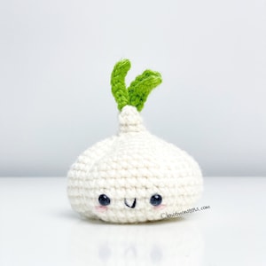 May include: A white crocheted garlic with a green stem and a smiling face. The garlic is sitting on a white surface.