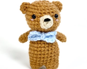 May include: A small brown crocheted teddy bear wearing a light blue bow tie. The bear has black eyes and a white nose. The bear is attached to a silver keychain.
