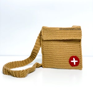 May include: A crocheted tan crossbody bag with a red circle and white plus sign on the front. The bag has a long strap and a flap closure. ©knotmonsters.com