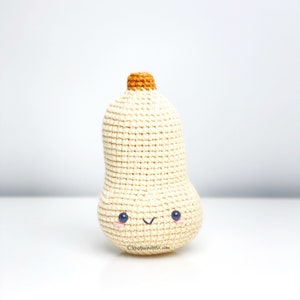 May include: A crocheted butternut squash with a smiling face. The squash is cream colored with a brown stem and black eyes.