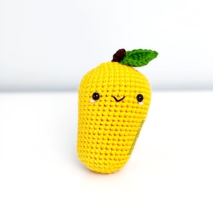 May include: A handmade, yellow crochet mango with a smiling face, black eyes, and a small, curved mouth. It has a brown stem and a green leaf. The mango is approximately 4 inches tall.