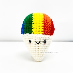 May include: A crocheted rainbow snow cone with a white cone and a cute face. The snow cone is made with green, yellow, orange, and red yarn.