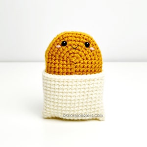 May include: A crocheted stuffed toy shaped like a potato with a smiling face. The potato is sitting in a white crocheted container.