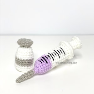 Puede incluir: Un conjunto de jeringa y vial de ganchillo, la jeringa es blanca con una franja morada y marcas negras, el vial es blanco con una franja gris.  ©knotmonsters.com