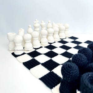 May include: A crocheted chess set with white and black pieces arranged on a black and white checkered board. The white pieces are in the foreground, with the black pieces in the background. The chess board is made of a soft, textured material.