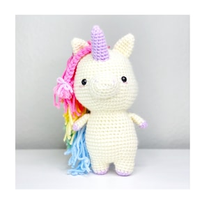 Peut inclure: Une licorne au crochet faite à la main, de couleur blanc cassé, avec une corne lavande, une crinière rose et arc-en-ciel, et une queue bleue. La licorne a de petits yeux noirs et des accents violets sur les sabots et les oreilles. Ce jouet est parfait pour les enfants.