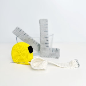 May include: A yellow crocheted tape measure with a black eye, a white crocheted tape, and two grey crocheted rulers in the background.