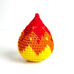 May include: A handmade crochet fire design in shades of red, orange, and yellow. The flame-shaped item is crocheted with a textured stitch, and the base is yellow, transitioning to orange and red at the top. The website address "knotmonsters.com" is visible.
