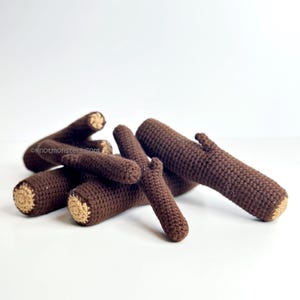 Op de afbeelding: Een verzameling bruine gehaakte boomstammen met bruine uiteinden ligt op een witte ondergrond. De boomstammen variëren in grootte en vorm, sommige met kleine takachtige uitsteeksels. De textuur van het haken is zichtbaar.