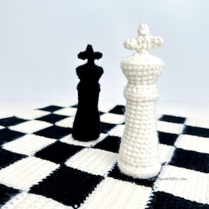 May include: A black and white crochet chess set featuring a black king and a white king. The chess pieces are on a black and white checkered board. The chess pieces are handmade and have a textured appearance.