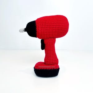 Może przedstawiać: Czerwono-czarna szydełkowa wiertarka ze srebrnym wiertłem. Wiertarka ma czarny uchwyt i podstawę. Adres strony internetowej "knotmonsters.com" jest widoczny na podstawie.